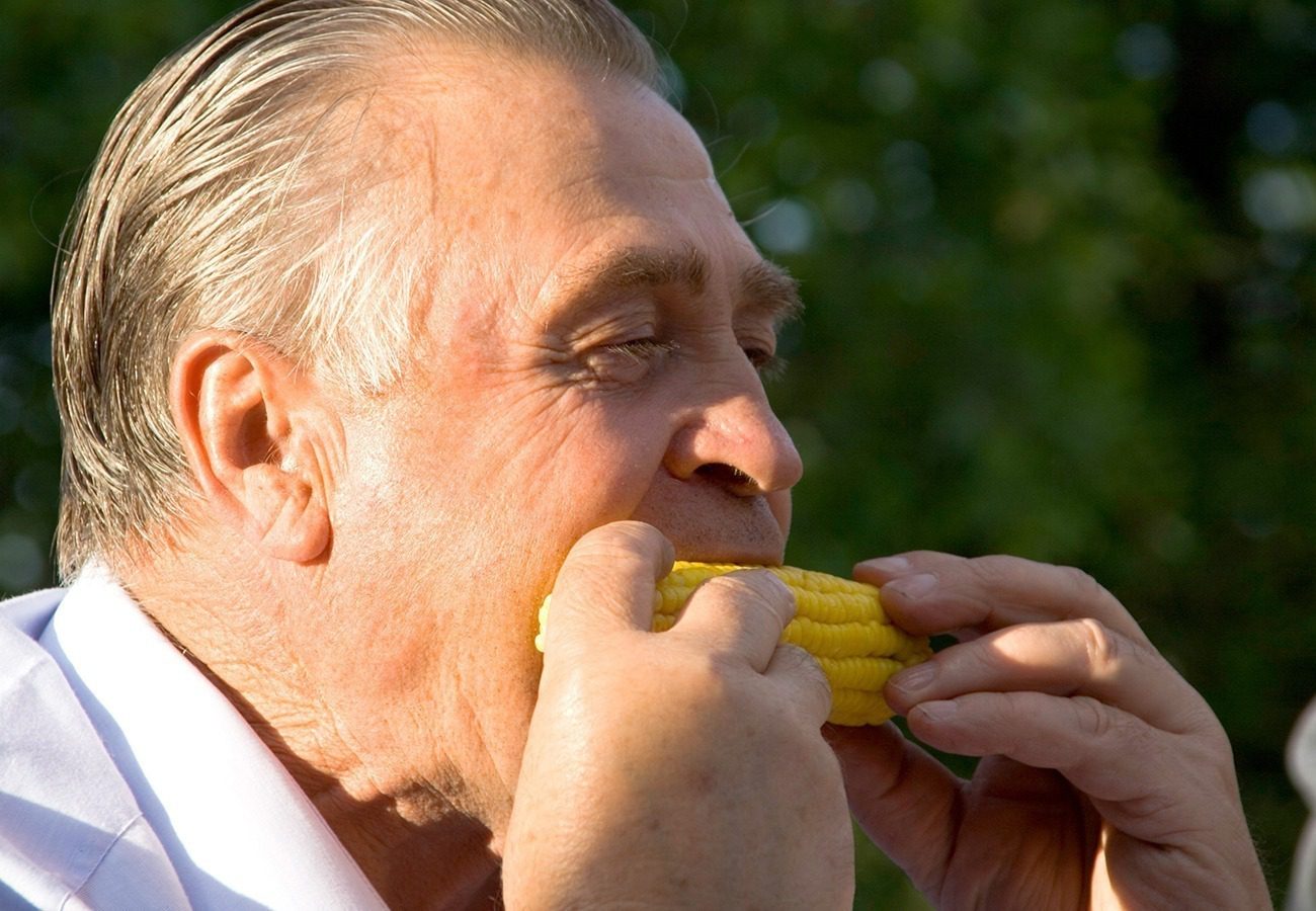 An older man with gray hair is eating corn on the cob outdoors, holding it with both hands. The background is blurred greenery, suggesting a sunny day.