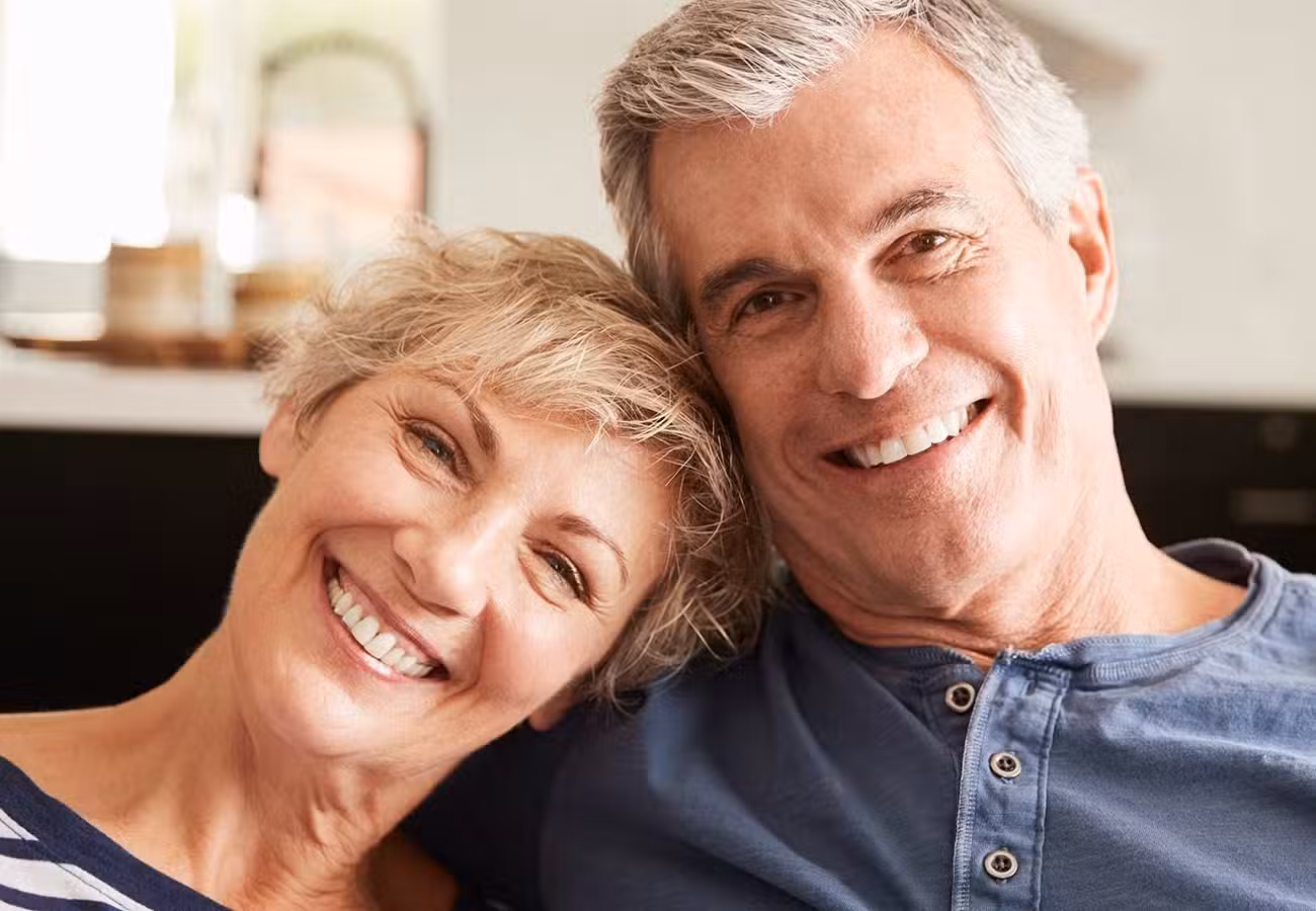 An older couple smiling and sitting closely together indoors, both appearing happy and relaxed. The woman is leaning her head on the mans shoulder. They have short gray hair and are casually dressed.