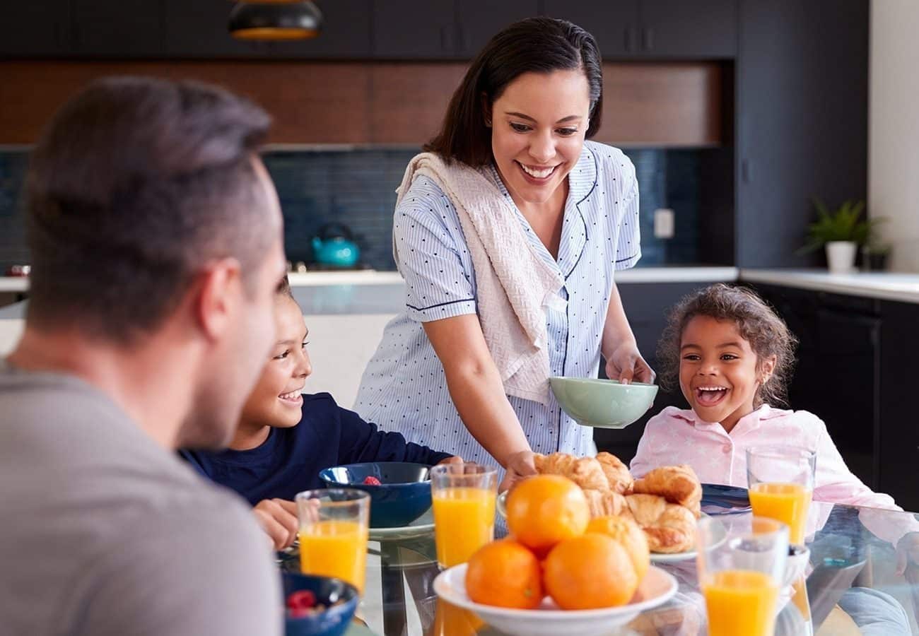 A woman serves breakfast to two smiling children and a man at a kitchen table set with pastries, oranges, berries, and glasses of orange juice.