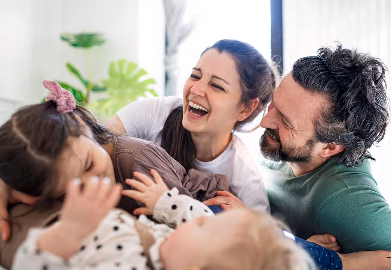 A happy family of four, including two adults and two young children, laughing and playing together indoors, creating a joyful and loving atmosphere.