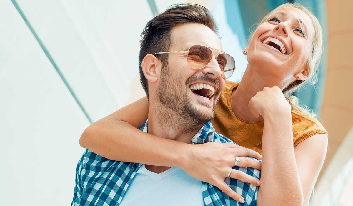 A smiling woman playfully hugs a laughing man from behind. Both appear happy and joyful, outdoors in bright sunlight. The man wears sunglasses and a plaid shirt; the woman has blonde hair and wears a sleeveless top.