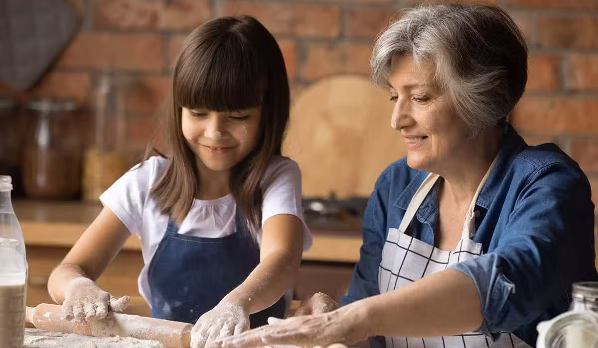 A smiling elderly woman and a young girl, both wearing aprons, roll out dough together on a floured surface in a cozy kitchen. The girl looks happy and focused, enjoying the baking activity.