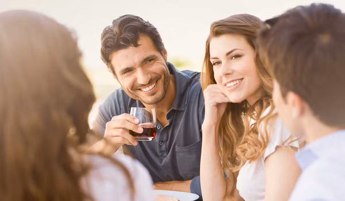 A group of four people sit together outdoors, smiling and talking. One man holds a glass of red wine. The atmosphere appears relaxed and friendly, with sunlight illuminating their faces.