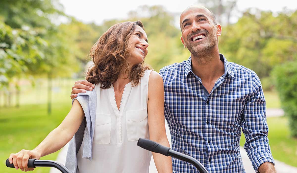 A smiling couple walks outdoors, holding bicycle handlebars. The woman has her arm around the man, and they look at each other happily. They are surrounded by greenery on a sunny day.
