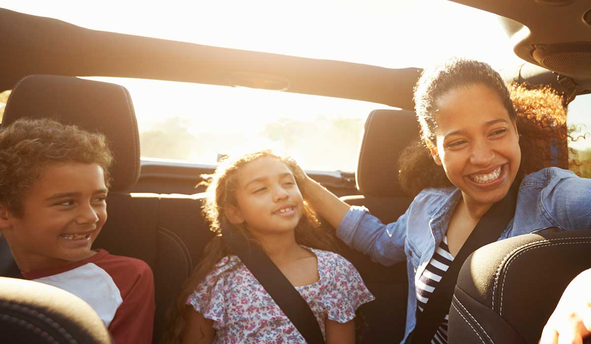 A woman smiles at two children sitting in the backseat of a car. The group appears happy, enjoying a sunny day with sunlight streaming in, and all are wearing seatbelts.